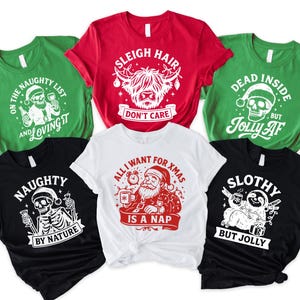 May include: A collection of Christmas-themed t-shirts in various colors, including green, red, black, and white. Each shirt features a unique graphic design with text, such as "Sleigh Hair Don't Care" and "Naughty by Nature".