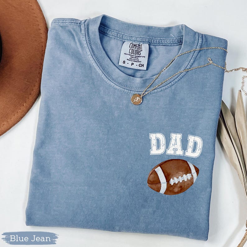 May include: A blue jean Comfort Colors t-shirt with the word "DAD" in white block letters and a watercolor football graphic. A gold necklace with a pendant is draped across the shirt.