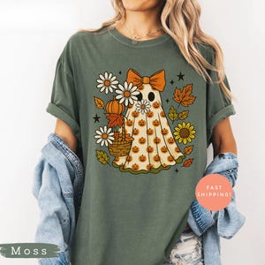 Puede incluir: Camiseta verde musgo con un diseño caprichoso de fantasma con un lazo naranja y una sábana estampada con calabazas. El diseño incluye margaritas, girasoles y hojas de otoño, perfecto para Halloween o el otoño.