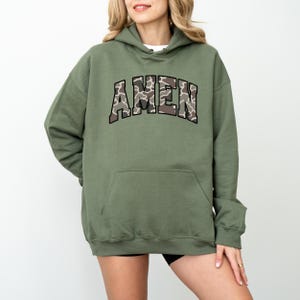 May include: An olive green hoodie with the word "AMEN" in a camouflage pattern across the chest. The hoodie has a drawstring hood and a large front pocket. The sleeves are long, and the material appears to be a soft, comfortable fabric.