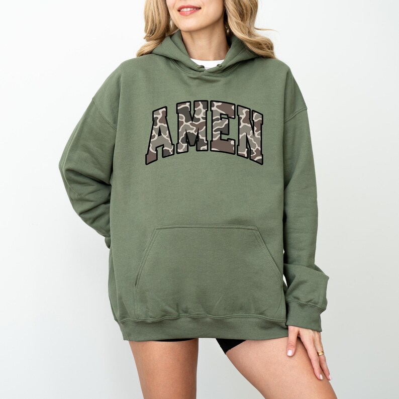 May include: An olive green hoodie with the word "AMEN" in a camouflage pattern across the chest. The hoodie has a drawstring hood and a large front pocket. The sleeves are long, and the material appears to be a soft, comfortable fabric.