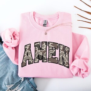 May include: A light pink sweatshirt with the word "AMEN" in a camouflage pattern across the front. The sweatshirt has long sleeves and a crew neck. A gold necklace with a small pendant is draped across the top.