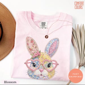 Pode incluir: Uma camiseta rosa claro Comfort Colors com um design de coelho em patchwork, óculos em forma de coração e detalhes florais. Um colar com um pingente está sobre a camiseta. A palavra "Blossom" está na parte inferior.