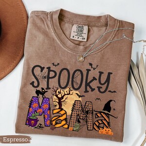 May include: Espresso-colored t-shirt with the words "Spooky Mama" in a Halloween-themed design. The design includes bats, spiderwebs, pumpkins, and a witch's hat. The shirt has a crew neck and short sleeves. A silver necklace with a pendant is on the shirt.