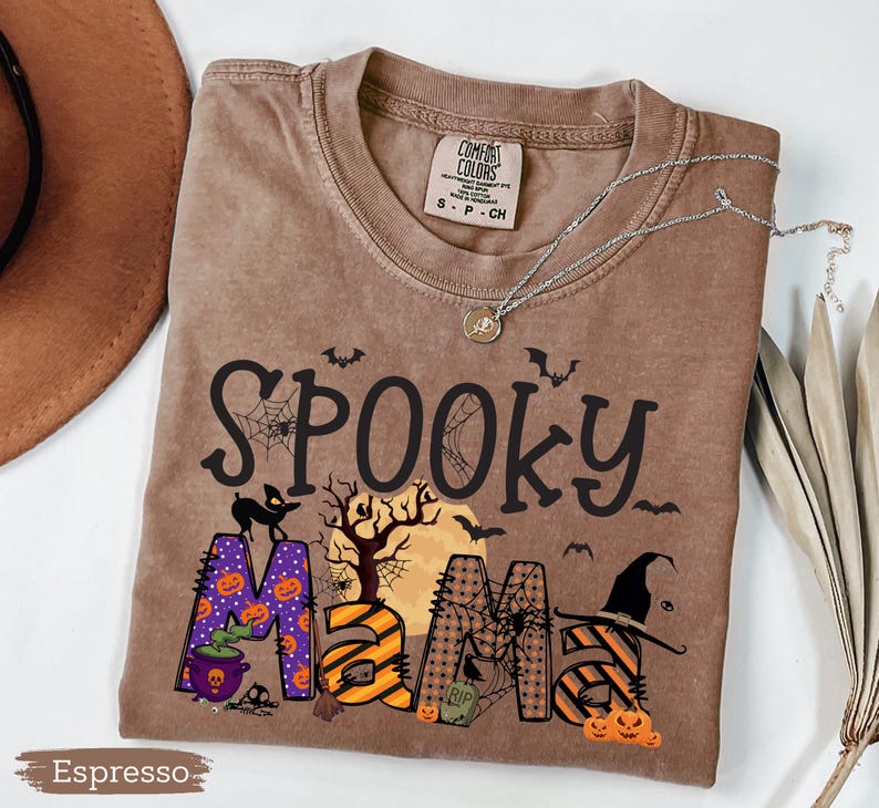 May include: Espresso-colored t-shirt with the words "Spooky Mama" in a Halloween-themed design. The design includes bats, spiderwebs, pumpkins, and a witch's hat. The shirt has a crew neck and short sleeves. A silver necklace with a pendant is on the shirt.