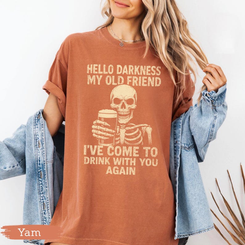 K&ouml;nnte beinhalten: Rostfarbenes T-Shirt mit dem Text "HELLO DARKNESS MY OLD FRIEND" &uuml;ber einem Skelett, das ein Bier h&auml;lt. Darunter steht der Text "I'VE COME TO DRINK WITH YOU AGAIN". Das Shirt hat kurze &Auml;rmel und eine lockere Passform.