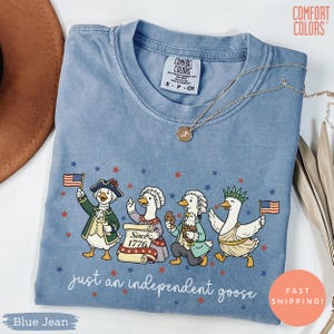 以下が含まれることがあります： 愛国的なガチョウのデザインが施された、ブルーデニムのComfort Colors Tシャツ。クルーネックと半袖です。グラフィックには、歴史的な衣装を着て旗やクッキーを持っているガチョウが描かれており、「just an independent goose」というテキストが添えられています。