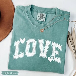 May include: A light green t-shirt with the word "LOVE" in white block letters and heart accents. The shirt is folded and has a silver necklace with a pendant. The tag reads "Comfort Colors".