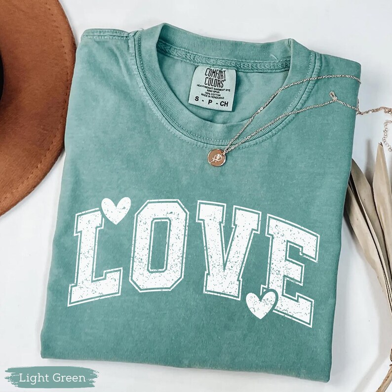 May include: A light green t-shirt with the word "LOVE" in white block letters and heart accents. The shirt is folded and has a silver necklace with a pendant. The tag reads "Comfort Colors".