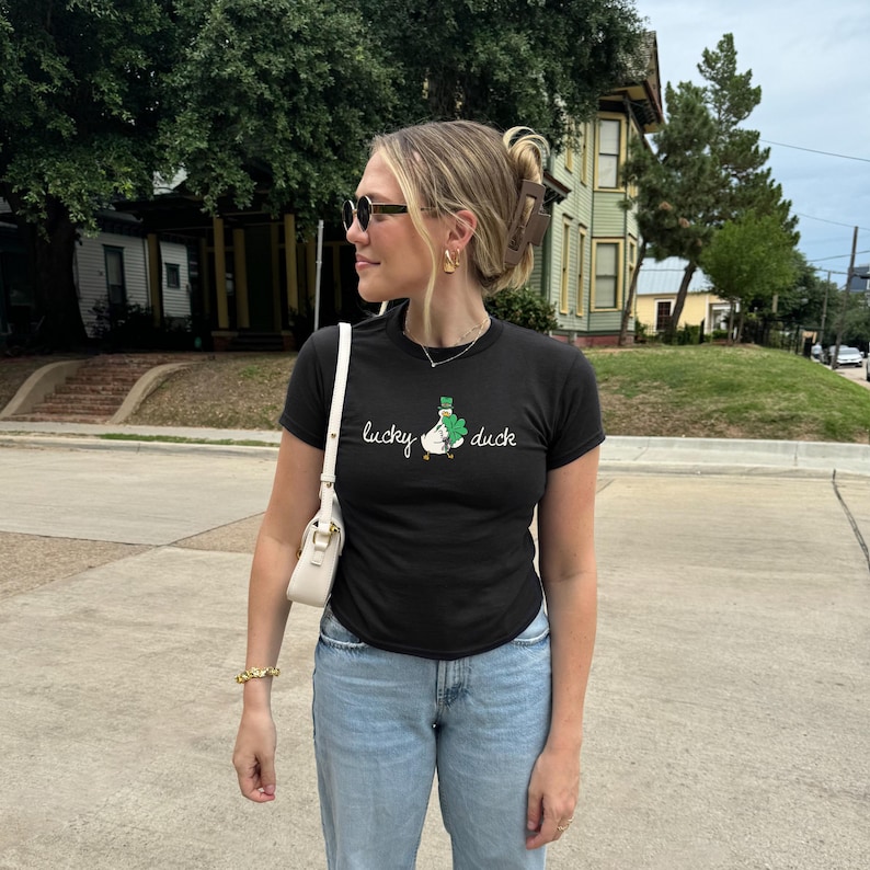 May include: Black t-shirt with the text "lucky duck" and a graphic of a duck wearing a shamrock. The shirt is worn with light blue jeans and a white handbag. The person is wearing sunglasses and gold jewellery.