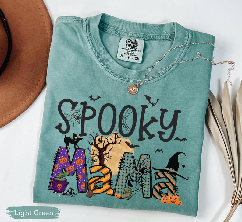 May include: Light green t-shirt with the words "Spooky Mama" in black, orange, and purple. The design includes bats, pumpkins, a witch's hat, and a cauldron. A silver necklace with a pendant is draped across the shirt.