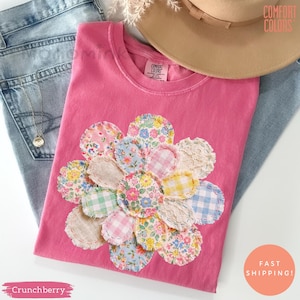 以下が含まれることがあります： 様々な布パッチで作られた、大きなカラフルな花の模様が特徴のピンク色のTシャツ。 Tシャツはクルーネックで、タグには「Comfort Colors」の文字が見えます。 左下には「Crunchberry」の文字があります。