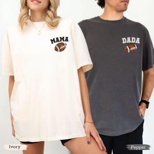 May include: Two short-sleeved t-shirts, one ivory and one pepper, featuring the words "MAMA" and "DADA" above a football graphic. The ivory shirt has a slightly oversized fit. The pepper shirt has a more fitted style.