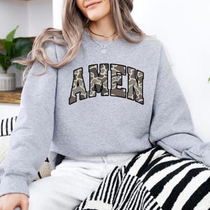 May include: A light gray sweatshirt with the word "AMEN" in a camouflage pattern across the chest. The sweatshirt has long sleeves and a crew neck. The design is in shades of brown, tan, and green.