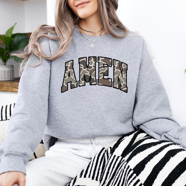 May include: A light gray sweatshirt with the word "AMEN" in a camouflage pattern across the chest. The sweatshirt has long sleeves and a crew neck. The design is in shades of brown, tan, and green.