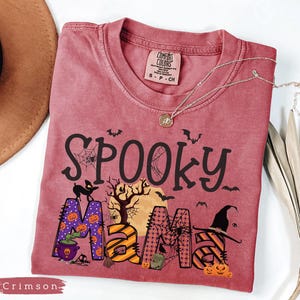 May include: Crimson-colored t-shirt with the words "Spooky Mama" in black, with Halloween-themed graphics. The "M" is purple with orange pumpkins. A silver necklace with a pendant is on the shirt.