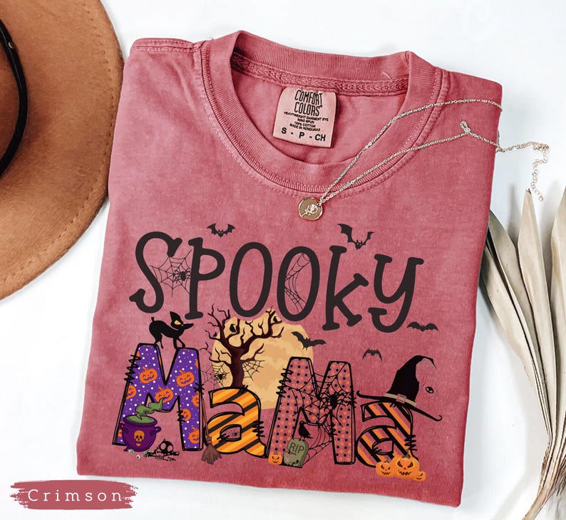 May include: Crimson-colored t-shirt with the words "Spooky Mama" in black, with Halloween-themed graphics. The "M" is purple with orange pumpkins. A silver necklace with a pendant is on the shirt.