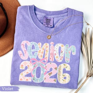 Może przedstawiać: Fioletowa koszulka Comfort Colors z napisem "Senior 2026" w patchworkowym wzorze. Wz&oacute;r zawiera kwiatowe i wzorzyste tkaniny w kolorze r&oacute;żowym, niebieskim i białym. Złoty naszyjnik z wisiorkiem jest zarzucony na koszulkę.