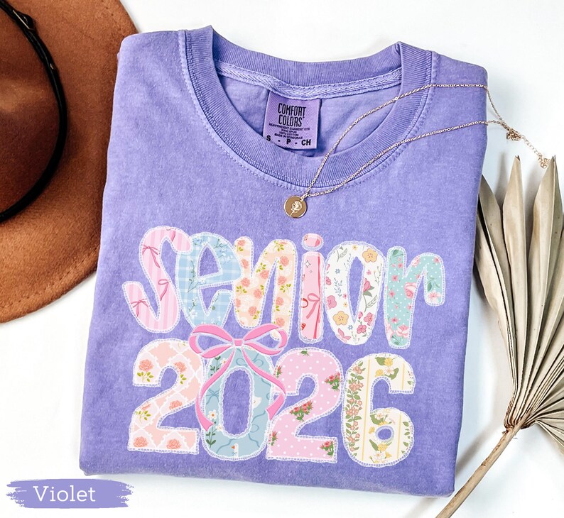 Może przedstawiać: Fioletowa koszulka Comfort Colors z napisem "Senior 2026" w patchworkowym wzorze. Wz&oacute;r zawiera kwiatowe i wzorzyste tkaniny w kolorze r&oacute;żowym, niebieskim i białym. Złoty naszyjnik z wisiorkiem jest zarzucony na koszulkę.