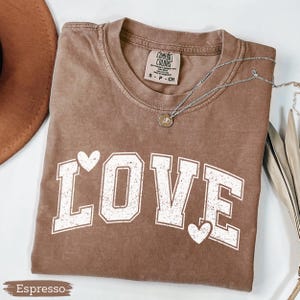 May include: Espresso-colored t-shirt with the word "LOVE" in white block letters and two white hearts. The shirt has a crew neck and short sleeves. A silver necklace with a small pendant is draped across the shirt.