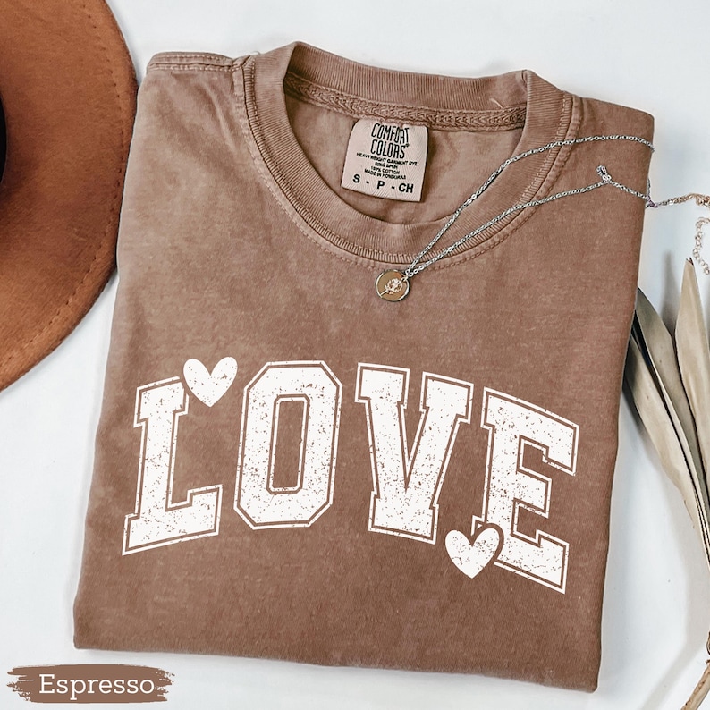 May include: Espresso-colored t-shirt with the word "LOVE" in white block letters and two white hearts. The shirt has a crew neck and short sleeves. A silver necklace with a small pendant is draped across the shirt.