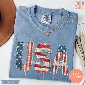 Puede incluir: Una camiseta azul vaquera Comfort Colors con la palabra "USA" aplicada en la parte delantera con un diseño de bandera estadounidense desgastada. Un collar de oro con un colgante está drapeado sobre la camisa. La camisa está doblada y expuesta sobre una superficie blanca.