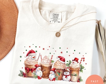 Puede incluir: Camiseta de color marfil con un diseño festivo de muñecos de nieve en tazas de café, con gorros de Papá Noel y bufandas. El diseño incluye detalles en rojo y verde, bastones de caramelo y estrellas. La camiseta tiene una etiqueta Comfort Colors.