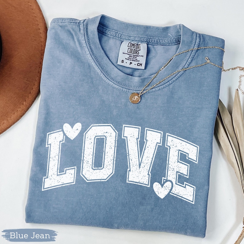 May include: A blue jean-colored t-shirt with the word "LOVE" in white block letters, with a heart above the "O" and below the "E". A gold necklace with a pendant rests on the shirt. The shirt has a Comfort Colors tag.