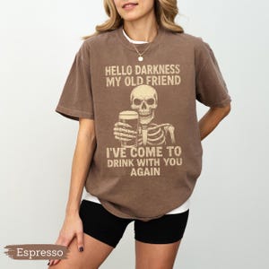 K&ouml;nnte beinhalten: Espressofarbenes T-Shirt mit einem Skelett, das ein Bier h&auml;lt, und dem Text "HELLO DARKNESS MY OLD FRIEND I'VE COME TO DRINK WITH YOU AGAIN". Das Shirt hat eine lockere Passform und wird mit schwarzen Shorts getragen.