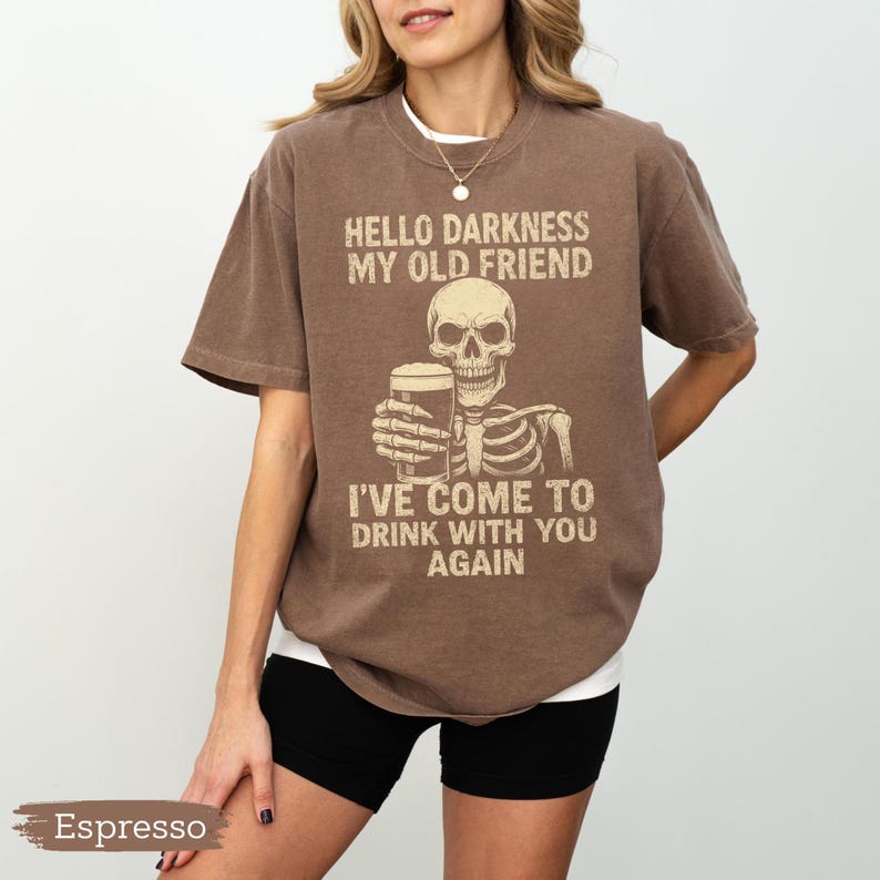 K&ouml;nnte beinhalten: Espressofarbenes T-Shirt mit einem Skelett, das ein Bier h&auml;lt, und dem Text "HELLO DARKNESS MY OLD FRIEND I'VE COME TO DRINK WITH YOU AGAIN". Das Shirt hat eine lockere Passform und wird mit schwarzen Shorts getragen.