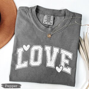 May include: A gray Comfort Colors t-shirt with the word "LOVE" in white block letters and heart accents. A silver necklace with a pendant is draped across the shirt. The shirt is folded on a white surface.