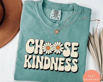 Puede incluir: Camiseta verde claro con las palabras "CHOOSE KINDNESS" en una fuente retro, con detalles de margaritas. La camiseta tiene cuello redondo y mangas cortas. Un collar plateado con un colgante descansa sobre la camiseta. La camiseta está etiquetada como "Comfort Colors".