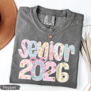 Może przedstawiać: Szara koszulka Comfort Colors z napisem "Senior 2026" w patchworkowym wzorze. Patchwork zawiera r&oacute;żowe, niebieskie i kwiatowe wzory. Srebrny naszyjnik z wisiorkiem leży na koszulce.