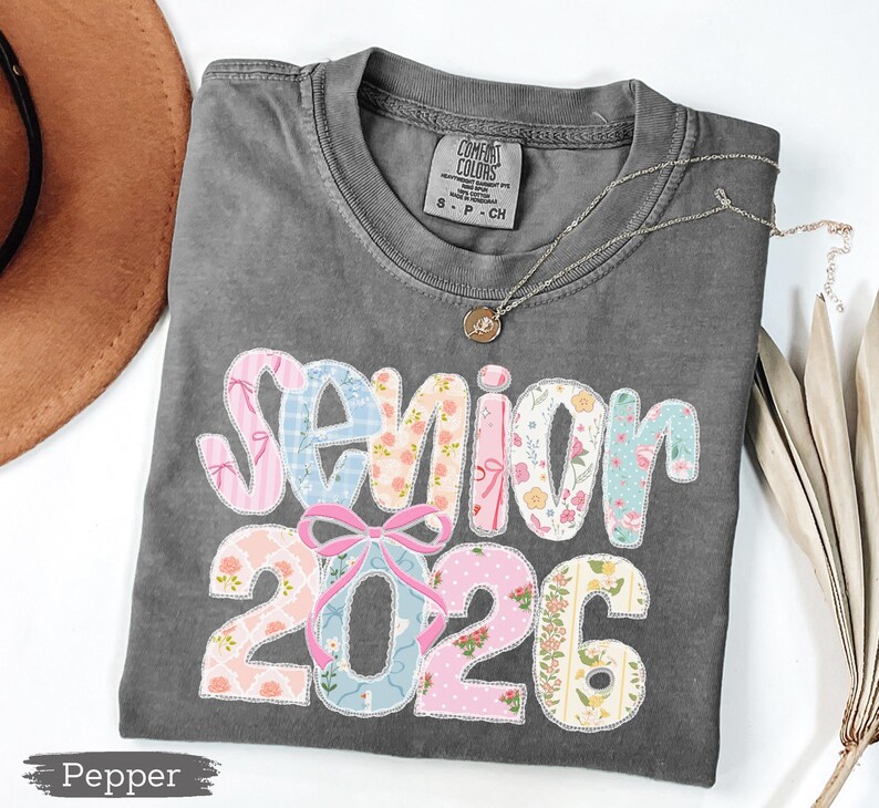 Może przedstawiać: Szara koszulka Comfort Colors z napisem "Senior 2026" w patchworkowym wzorze. Patchwork zawiera r&oacute;żowe, niebieskie i kwiatowe wzory. Srebrny naszyjnik z wisiorkiem leży na koszulce.