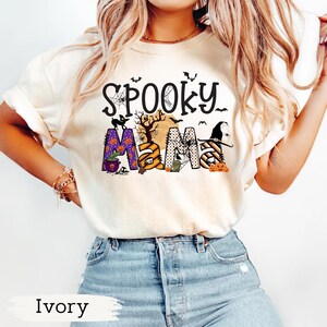 May include: Ivory-colored t-shirt with the words "Spooky Mama" in a Halloween-themed design. The design includes bats, a spiderweb, a witch hat, pumpkins, and the letters "MAMA" in various patterns and colors.