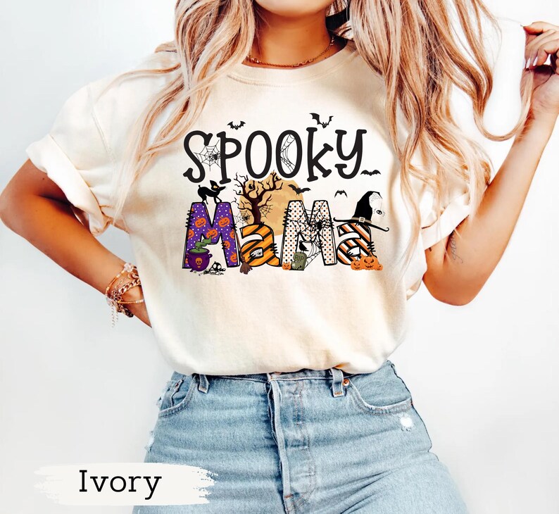 May include: Ivory-colored t-shirt with the words "Spooky Mama" in a Halloween-themed design. The design includes bats, a spiderweb, a witch hat, pumpkins, and the letters "MAMA" in various patterns and colors.