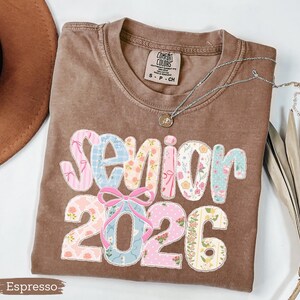Może przedstawiać: Koszulka w kolorze espresso z napisem "Senior 2026" w patchworkowym wzorze. Litery i cyfry są w pastelowych kolorach i kwiatowych wzorach. Srebrny naszyjnik z wisiorkiem jest zarzucony na koszulkę.