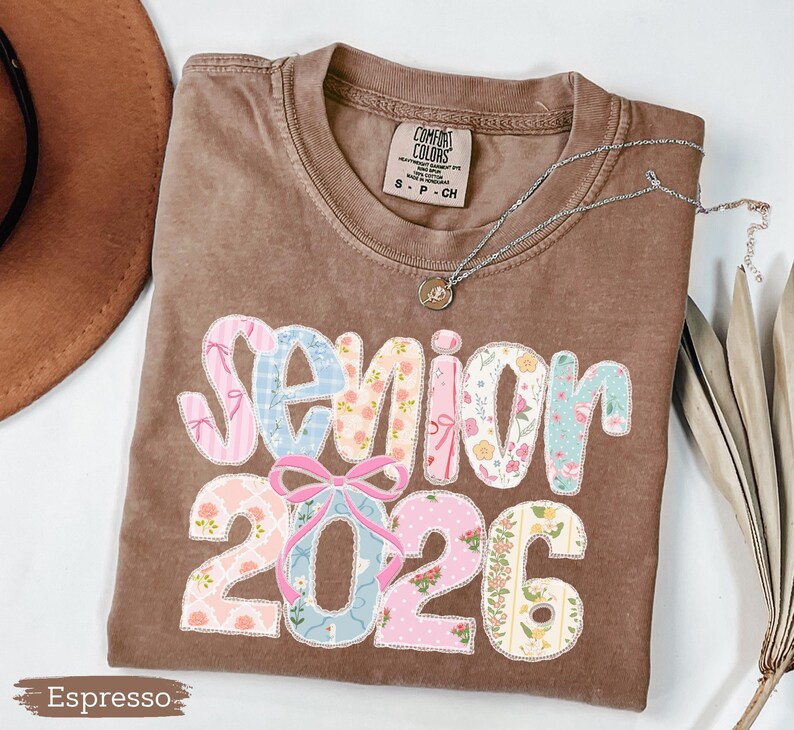 Może przedstawiać: Koszulka w kolorze espresso z napisem "Senior 2026" w patchworkowym wzorze. Litery i cyfry są w pastelowych kolorach i kwiatowych wzorach. Srebrny naszyjnik z wisiorkiem jest zarzucony na koszulkę.