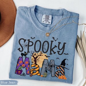 May include: Blue jean colored t-shirt with the words "Spooky Mama" in black, orange, and purple. The design includes bats, a black cat, pumpkins, a witch's hat, and a full moon. A gold necklace is draped across the shirt.