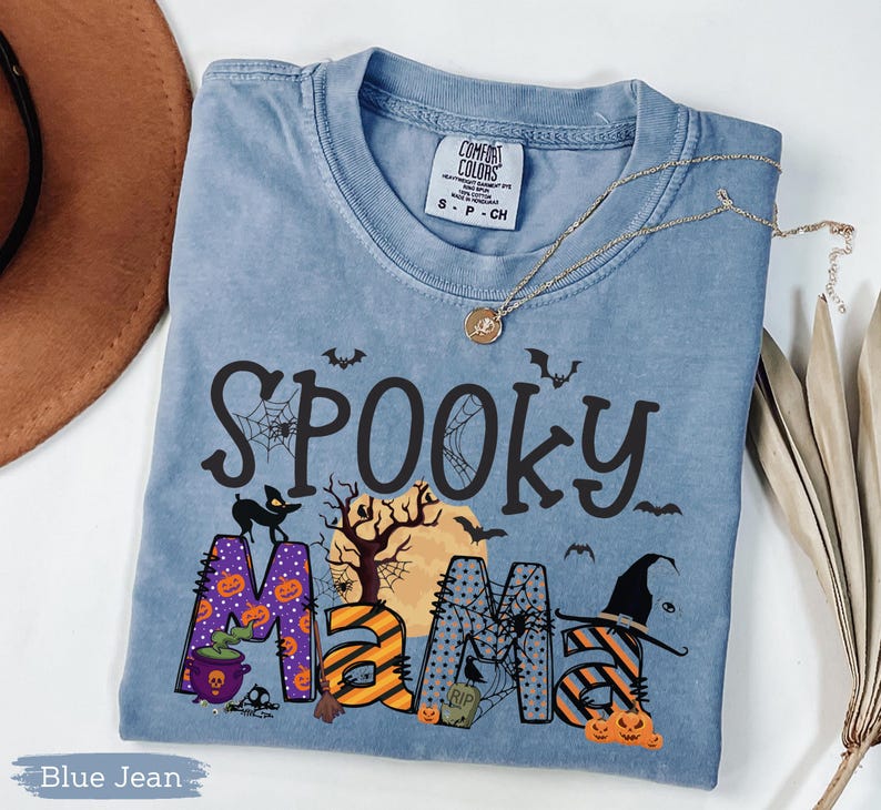 May include: Blue jean colored t-shirt with the words "Spooky Mama" in black, orange, and purple. The design includes bats, a black cat, pumpkins, a witch's hat, and a full moon. A gold necklace is draped across the shirt.