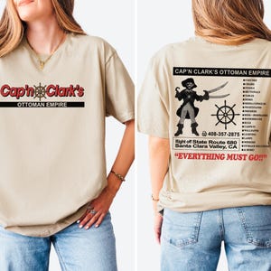 Peut inclure: Un t-shirt beige &agrave; manches courtes avec le texte rouge Cap'n Clark's Ottoman Empire sur le devant. Le dos pr&eacute;sente un graphique de pirate, une roue de navire, le texte Cap'n Clark's Ottoman Empire, une liste de meubles et Everything Must Go.