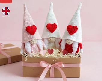 May include: Three crochet gnomes with tall, pointed hats and red heart accents sit in a brown box with decorative paper. The gnomes have white beards and are dressed in pink and red. A pink ribbon adorns the box. The text "VALENTINE HEART GNOME CROCHET PATTERN" is at the bottom.