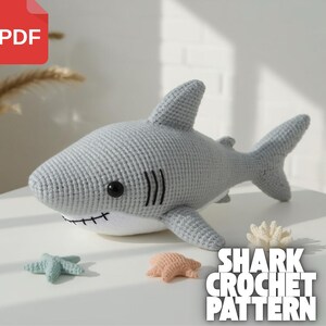 Puede incluir: Un juguete de tiburón gris tejido a ganchillo con ojos negros y vientre blanco. El tiburón tiene una boca cosida y branquias negras. Dos estrellas de mar y un coral tejidos a ganchillo están junto al tiburón. El texto "SHARK CROCHET PATTERN" está en la esquina inferior derecha.