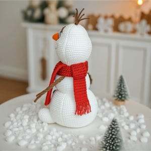 May include: A crocheted snowman figure with a red scarf, brown twig arms, and a carrot nose. The snowman has brown twig hair and is sitting on a white surface with faux snow. The snowman is a winter holiday decoration.