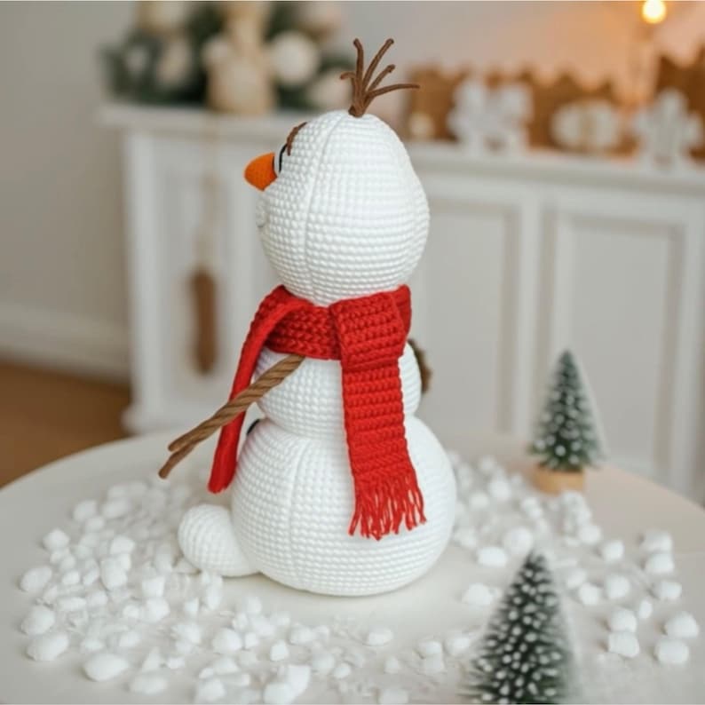 May include: A crocheted snowman figure with a red scarf, brown twig arms, and a carrot nose. The snowman has brown twig hair and is sitting on a white surface with faux snow. The snowman is a winter holiday decoration.