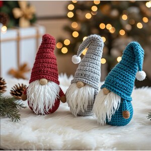 May include: Three handmade crochet gnomes in red, gray, and blue. Each gnome has a long, pointed hat with a white pom-pom, a white beard, and a small, round nose. They are displayed on a fluffy white surface, with Christmas lights in the background.