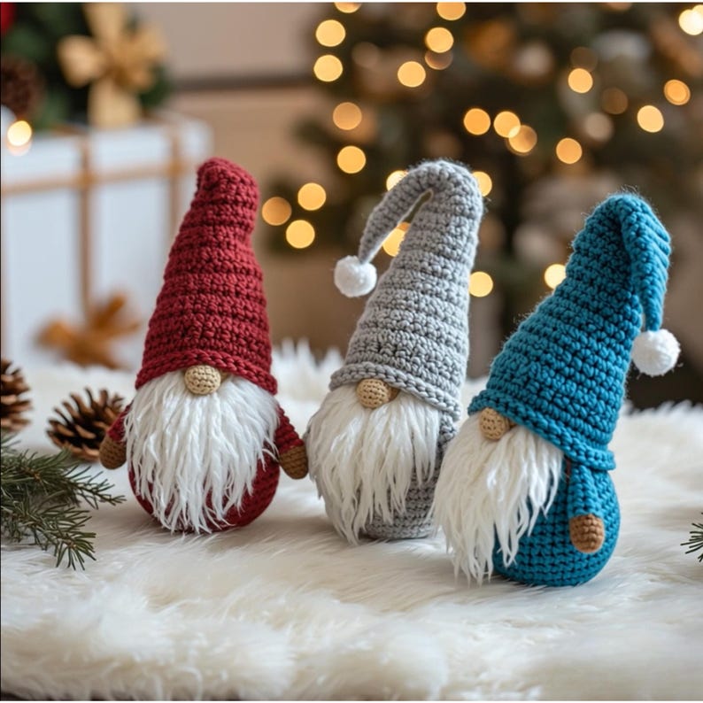 May include: Three handmade crochet gnomes in red, gray, and blue. Each gnome has a long, pointed hat with a white pom-pom, a white beard, and a small, round nose. They are displayed on a fluffy white surface, with Christmas lights in the background.