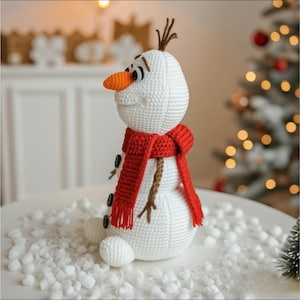 May include: A crocheted snowman with a carrot nose, black button eyes, and a red scarf. The snowman has brown twig hair and is sitting on a white surface with faux snow. A Christmas tree is visible in the background.