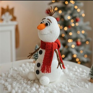 May include: A crocheted snowman with a carrot nose, black button eyes, and a red scarf. The snowman has twig arms and a smiling face. It is sitting on a white surface with faux snow, with a Christmas tree in the background.