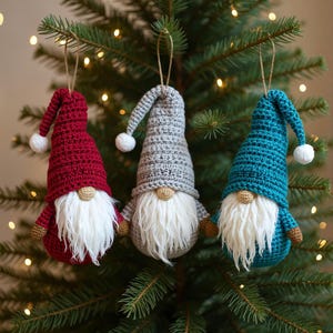 May include: Three handmade crochet gnome ornaments in red, gray, and teal blue, each with a white beard, a small tan nose, and a matching hat with a white pom-pom. The ornaments hang from a Christmas tree, creating a festive holiday display.